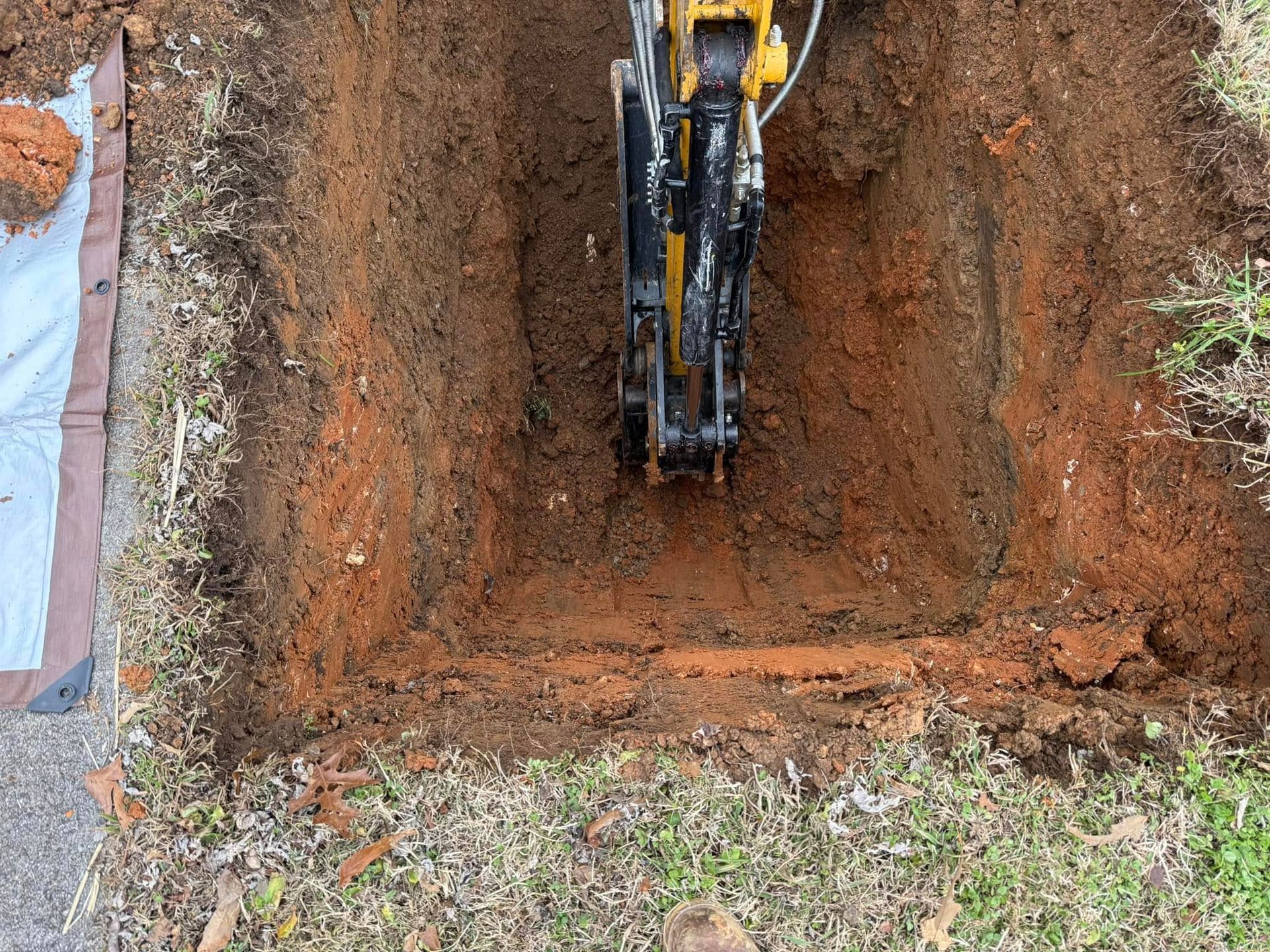 Excavator digging a rectangular trench in reddish soil, viewed from above.