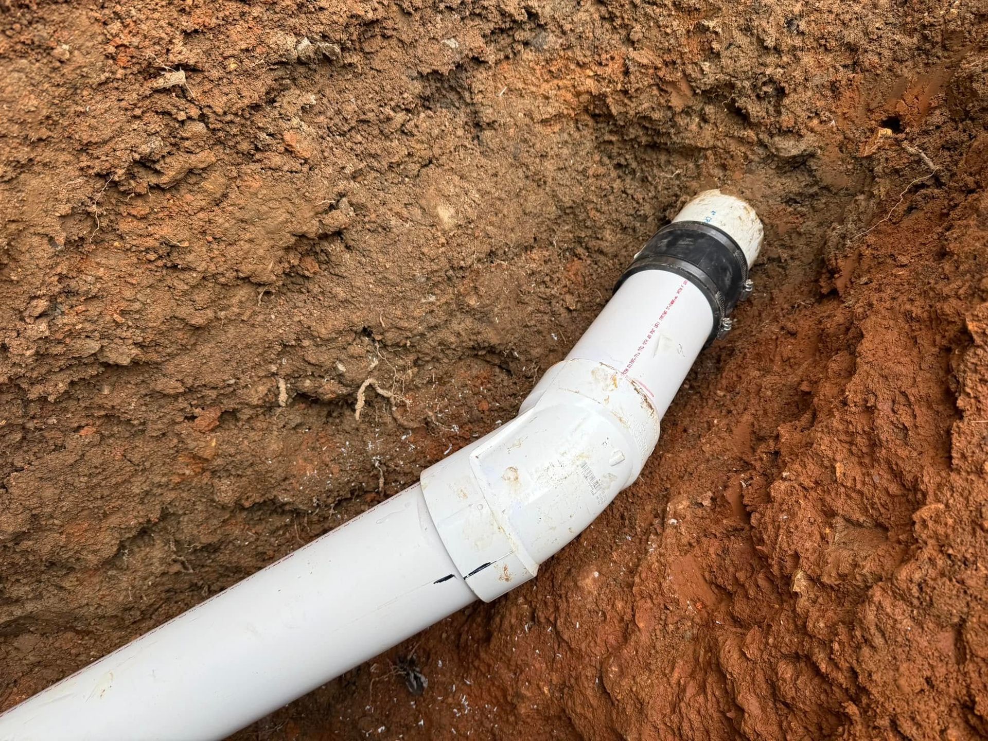 White PVC pipe elbow in a dirt trench with a black fitting at the end