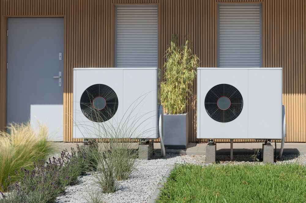 Two white heat pump units outside a house with wooden siding, next to plants and gravel.