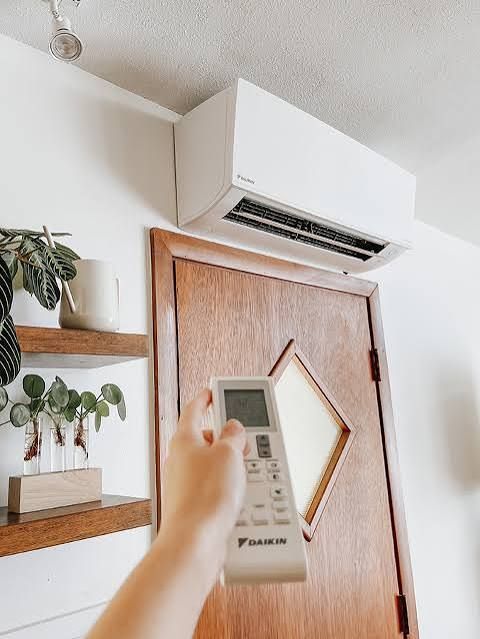 Hand holding remote, aiming at a white ductless air conditioner above a wooden door, indoors.