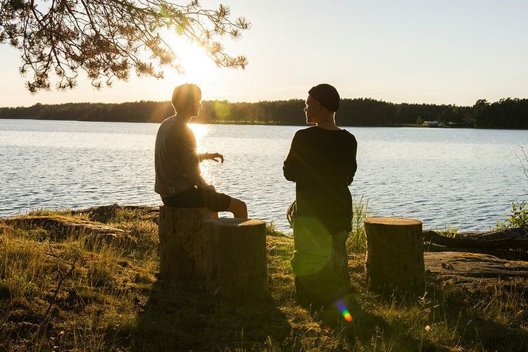 Two people sit on tree stumps by a lake, silhouetted against the setting sun.