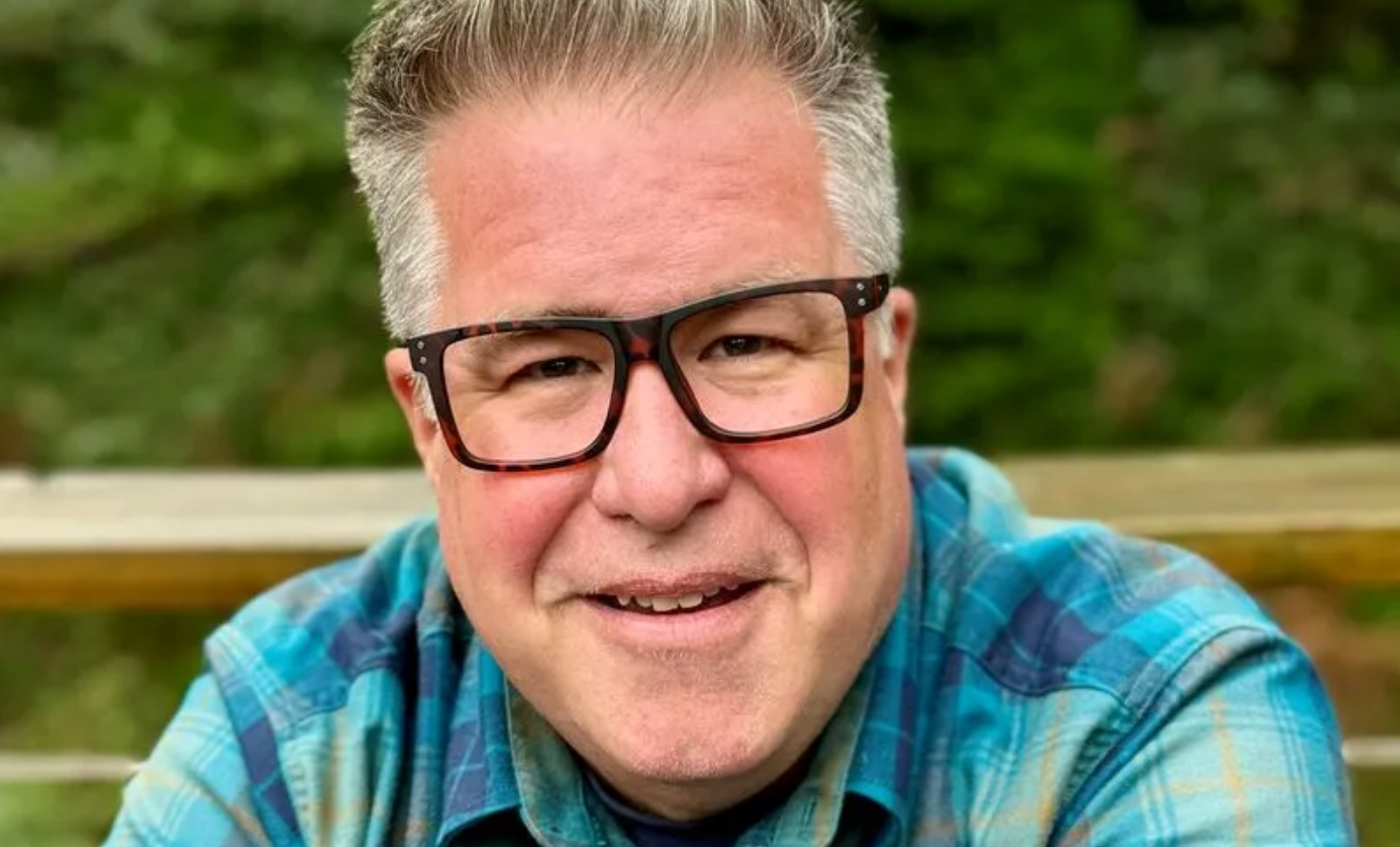 A person with gray hair and glasses wearing a blue plaid shirt, smiling warmly against a blurred outdoor background.
