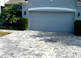 Grey paver driveway leading to a gray garage door. Green shrubs flank the entrance.