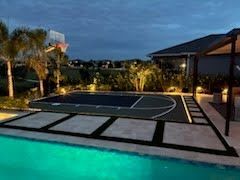 Backyard basketball court beside a lit pool at dusk, with palm trees and a covered patio.
