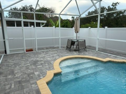 Screened-in backyard pool with gray pavers, white fence, and patio chairs under an umbrella