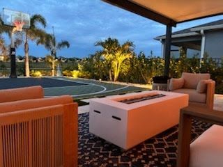 Modern patio seating beside a lit pool at dusk, with palm trees and a covered terrace.
