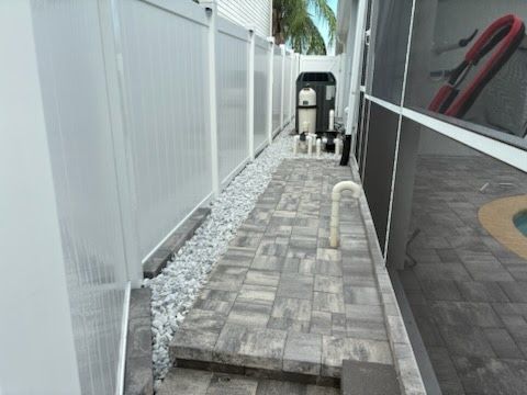 Narrow side yard with paver path, white fence, gravel border, and patio chairs in the background