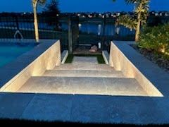 Modern outdoor pool steps descending to a waterfront deck at dusk, flanked by lit palm trees.