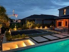 Modern backyard at dusk with a lit swimming pool, patio seating, and a house with warm interior lights