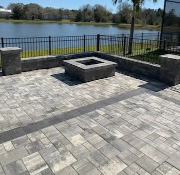 Patio with gray pavers, built-in fire pit, low wall, black fence, and view of a lake.