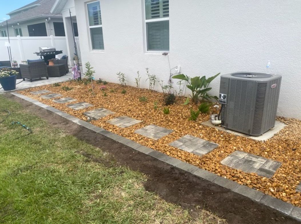 A backyard with a stone walkway, landscaping, and an AC unit next to a white house. The ground is covered with gravel