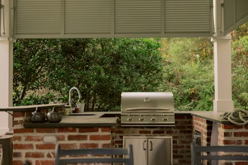 Outdoor kitchen with a brick structure, grill, sink, and lush green trees in the background.