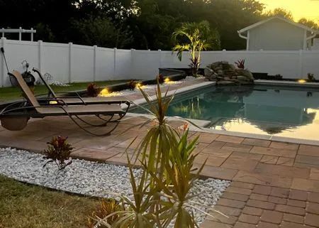 Swimming pool with brick patio, lounge chair, white fence, and landscaping.