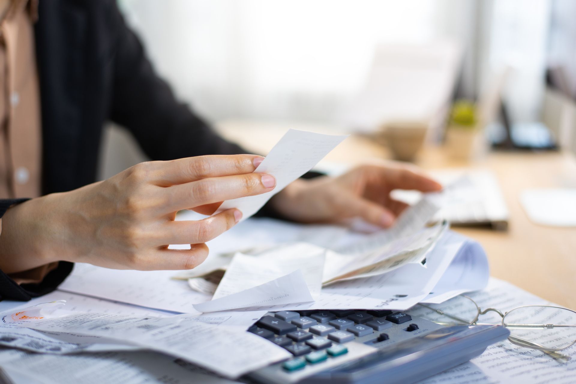 A woman is holding a piece of paper in front of a pile of papers and a calculator.