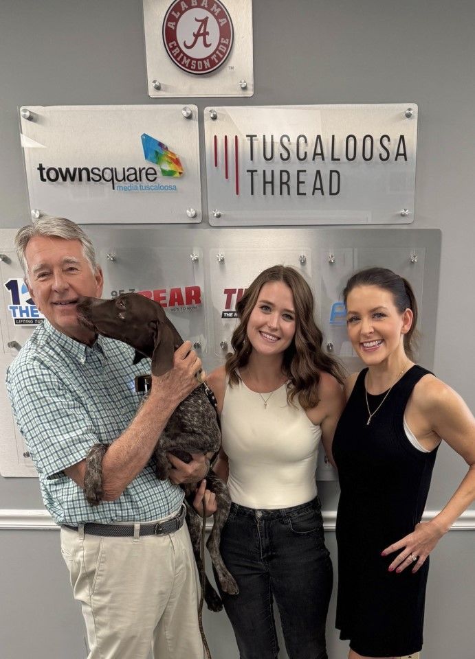Three people pose with a dog in front of a Tuscaloosa Thread sign and sponsor logos.
