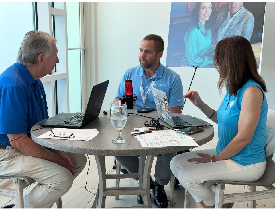 Three people in blue shirts seated around a table with a laptop in a bright meeting room
