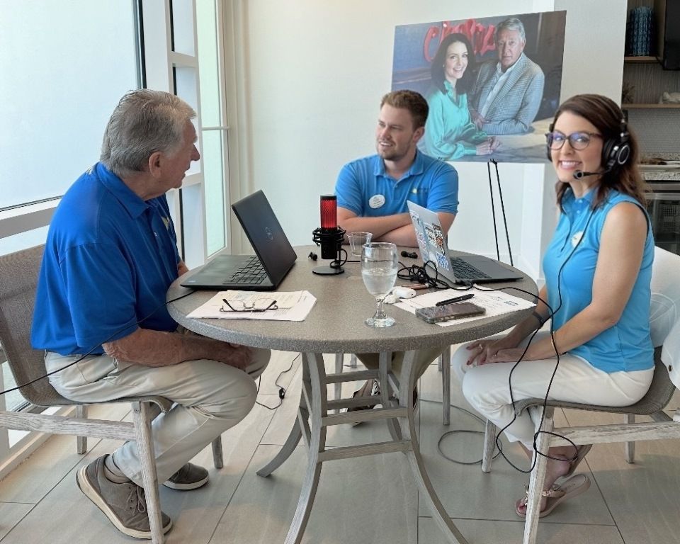 Three people in blue shirts sit at a table with laptops and headsets in a bright office.