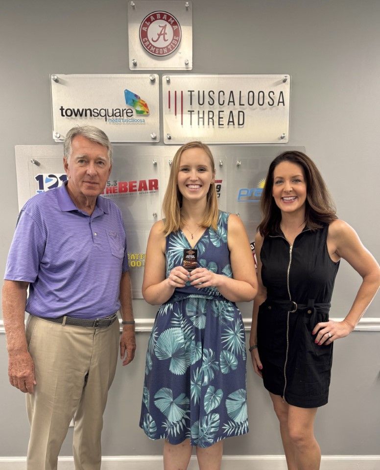 Three people posing indoors in front of business signs, one holding an award trophy.