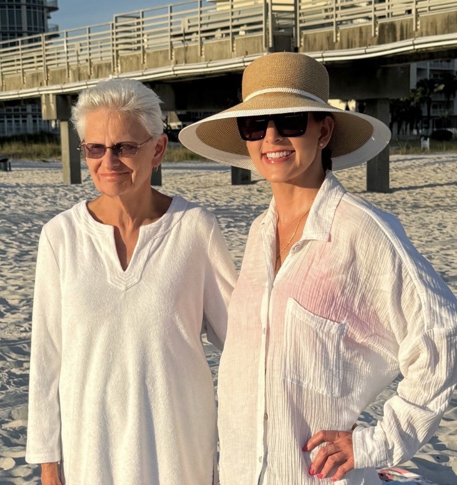 Two women in white cover-ups standing on a beach, one wearing a sunhat and sunglasses.