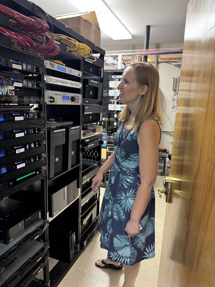 Woman standing in a server room, looking at stacked black network equipment and cables.