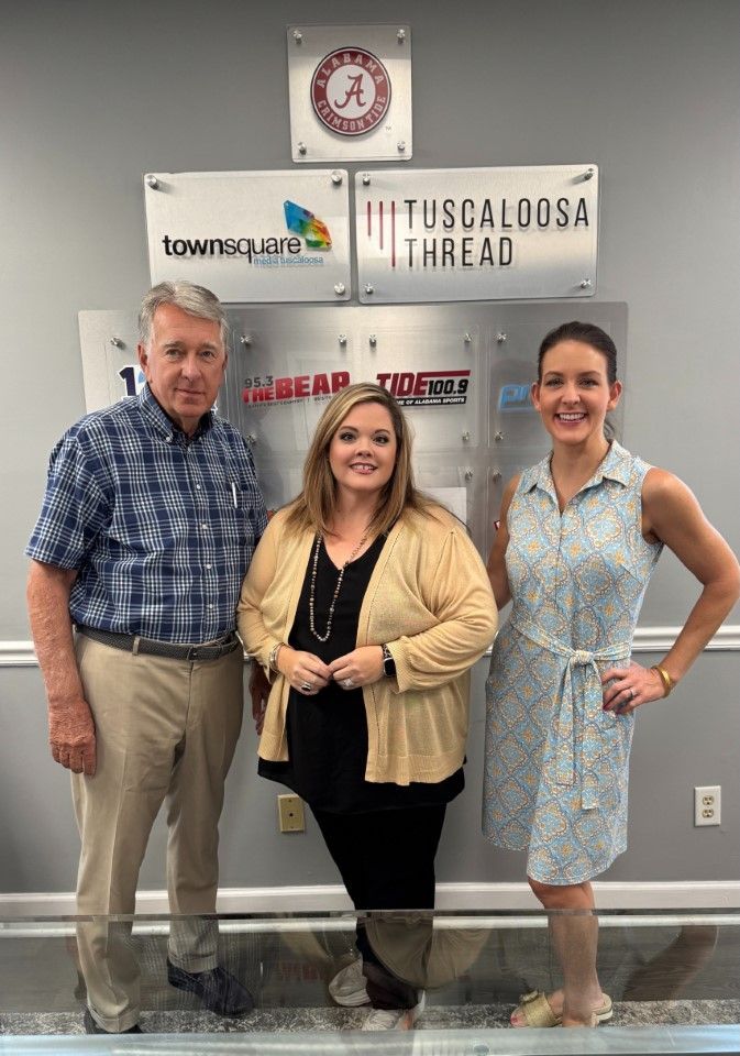 Three people posing indoors in front of a sponsor wall, smiling for a photo.