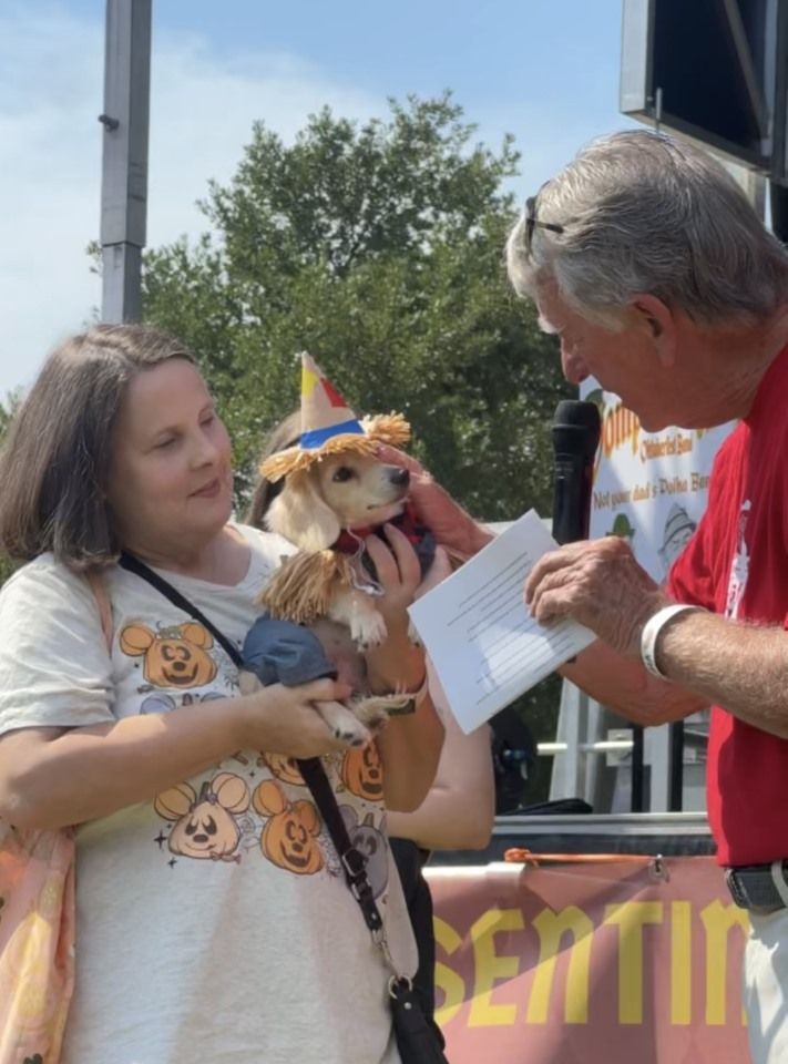 Woman holding a doll while a man in a red shirt reads from paper onstage outdoors