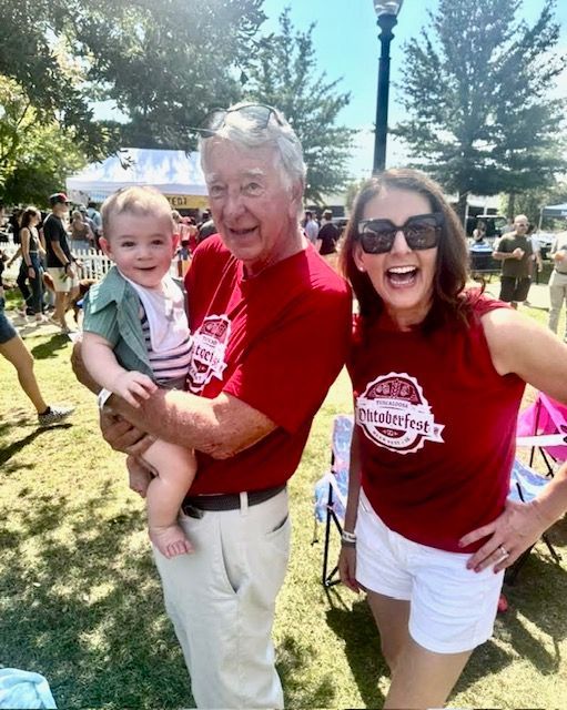 Three smiling people in red shirts posing outdoors at a sunny event, with a child being held.