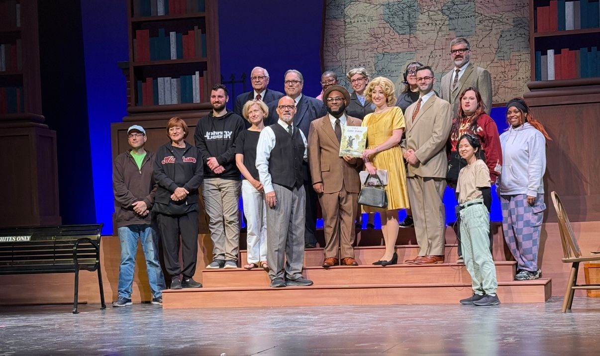 Group of people posing on a theater stage with bookshelves and warm lighting in the background