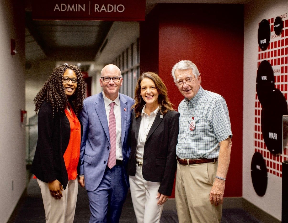 Four people posing in a hallway outside an admin radio room, smiling at the camera.