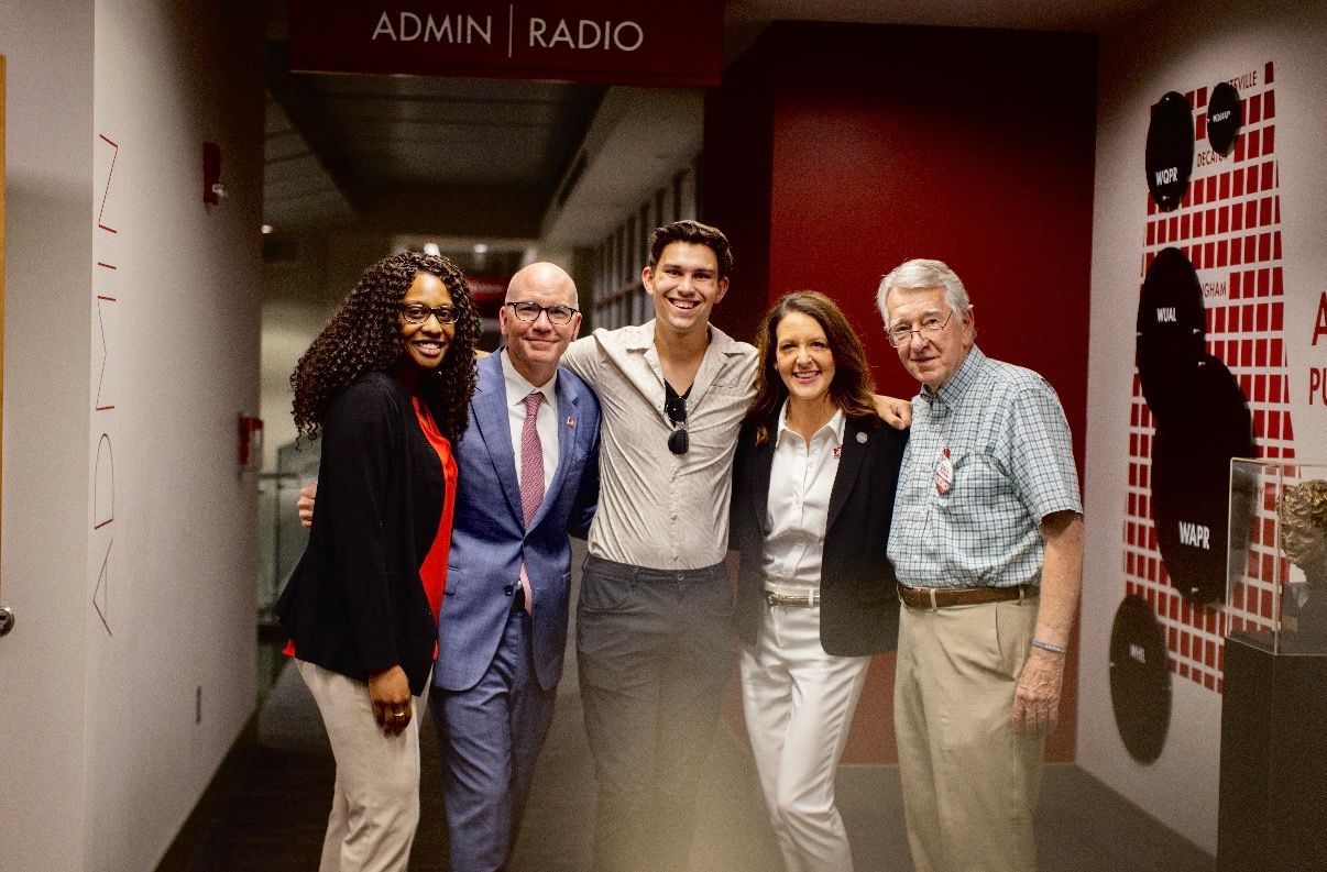 Five people posing in a hallway outside an “Admin Radio” sign