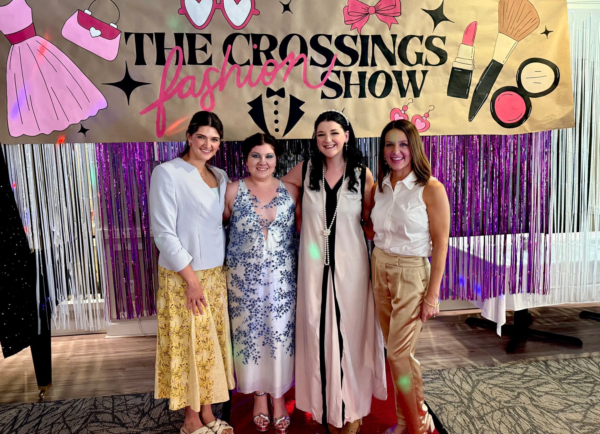 Four women posing in front of “The Crossings Fashion Show” backdrop with a purple fringe curtain