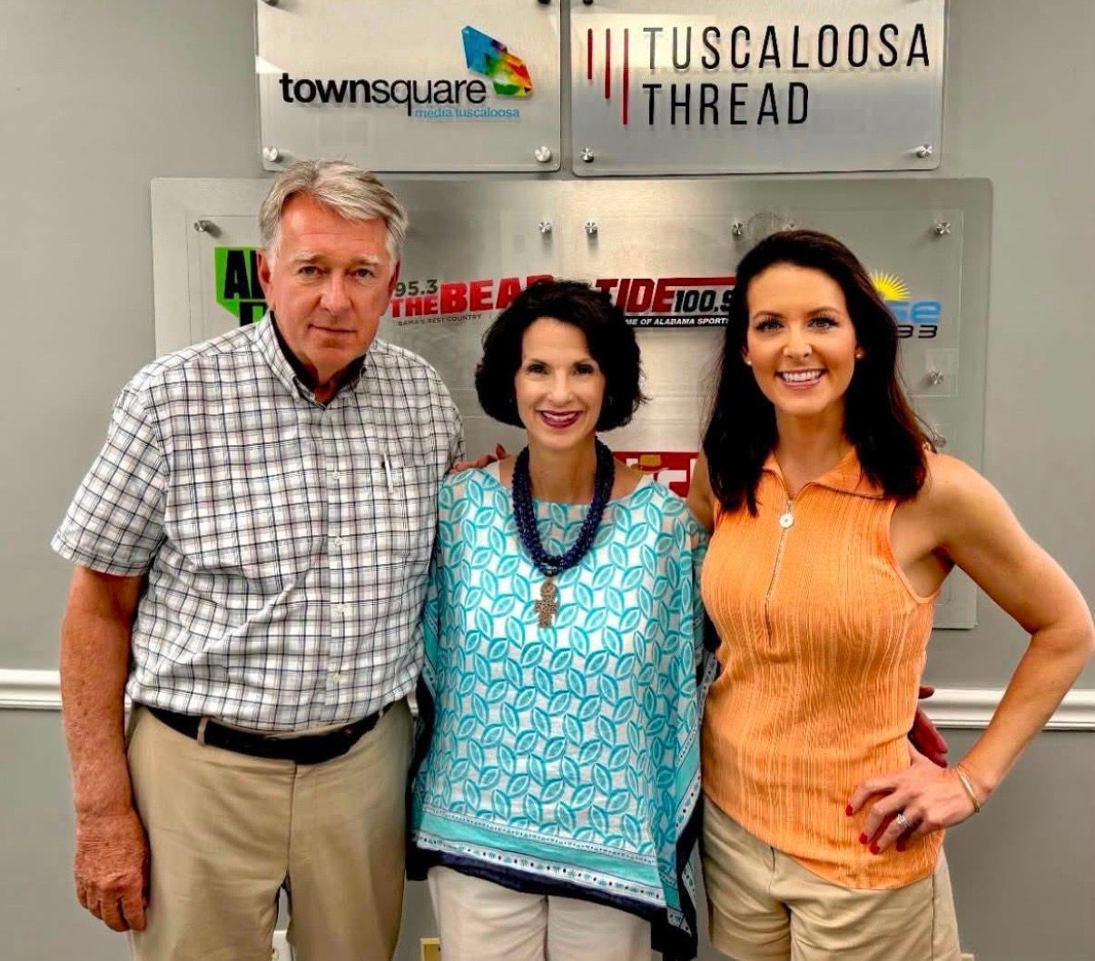 Three people posing in front of a Tuscaloosa Thread sign, smiling indoors.