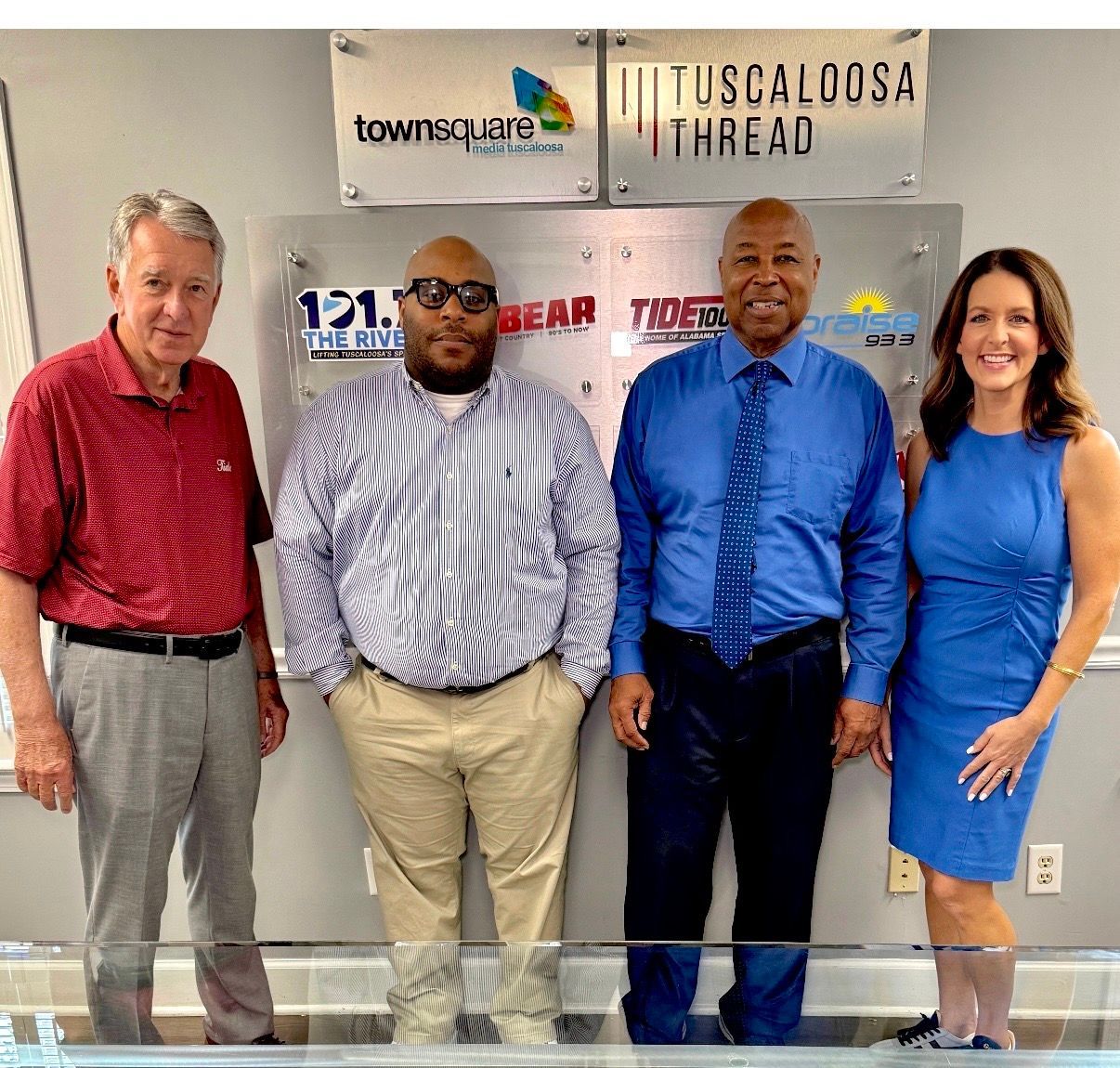 Four people posing indoors in front of Tuscaloosa Thread and townsquare logos.