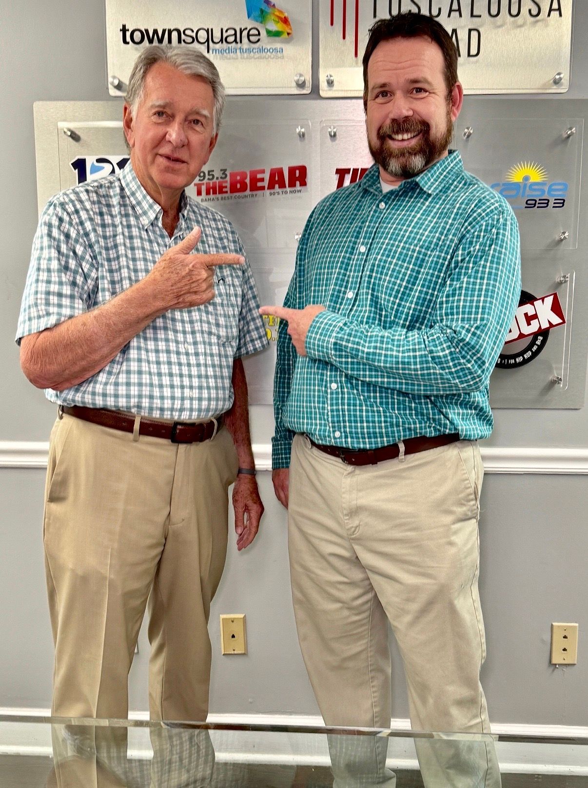 Two men in patterned shirts pointing at each other in a radio station studio