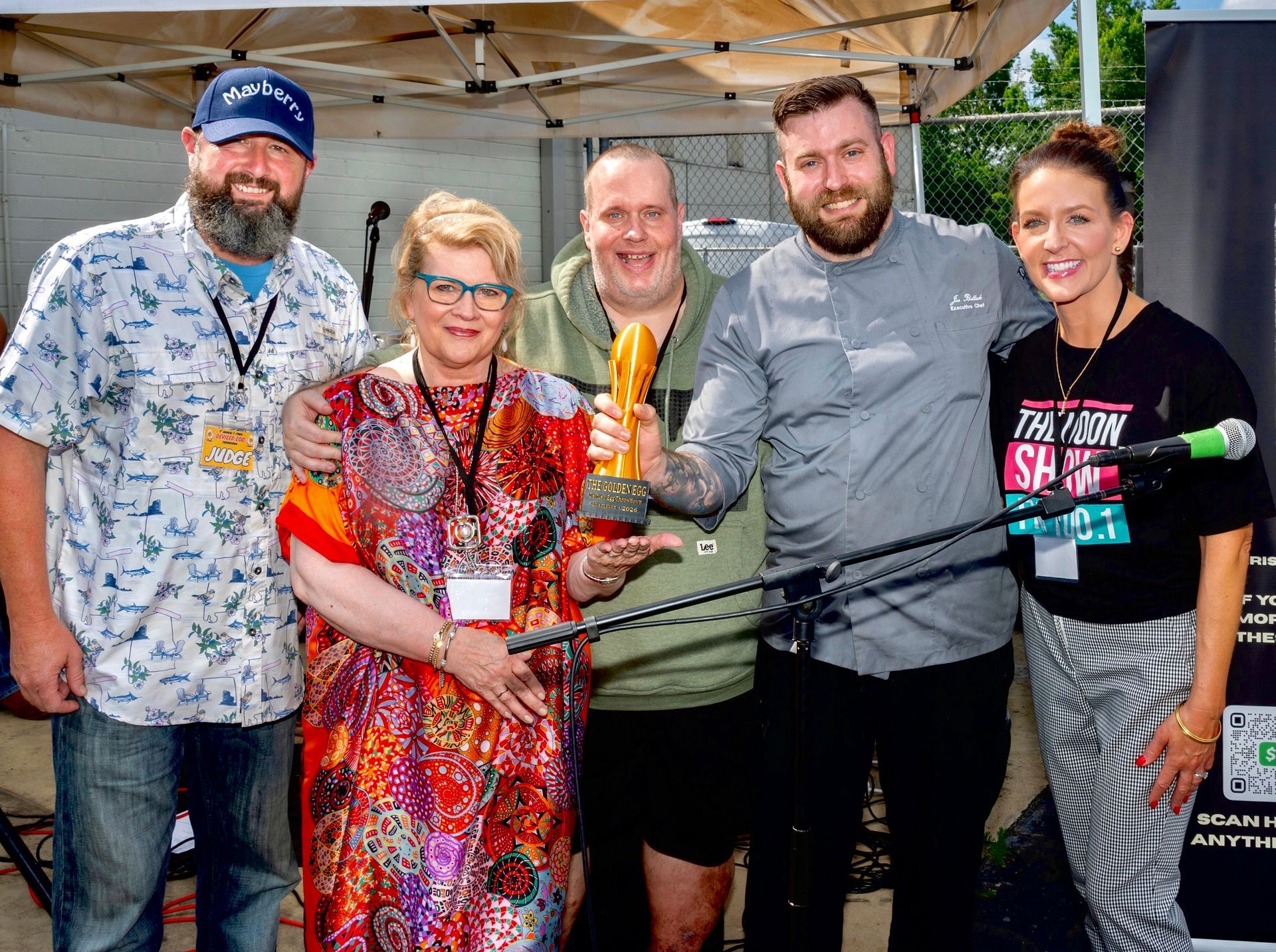 Five people posing under a canopy, smiling and holding ribbon-cutting scissors with a giant ceremonial key.