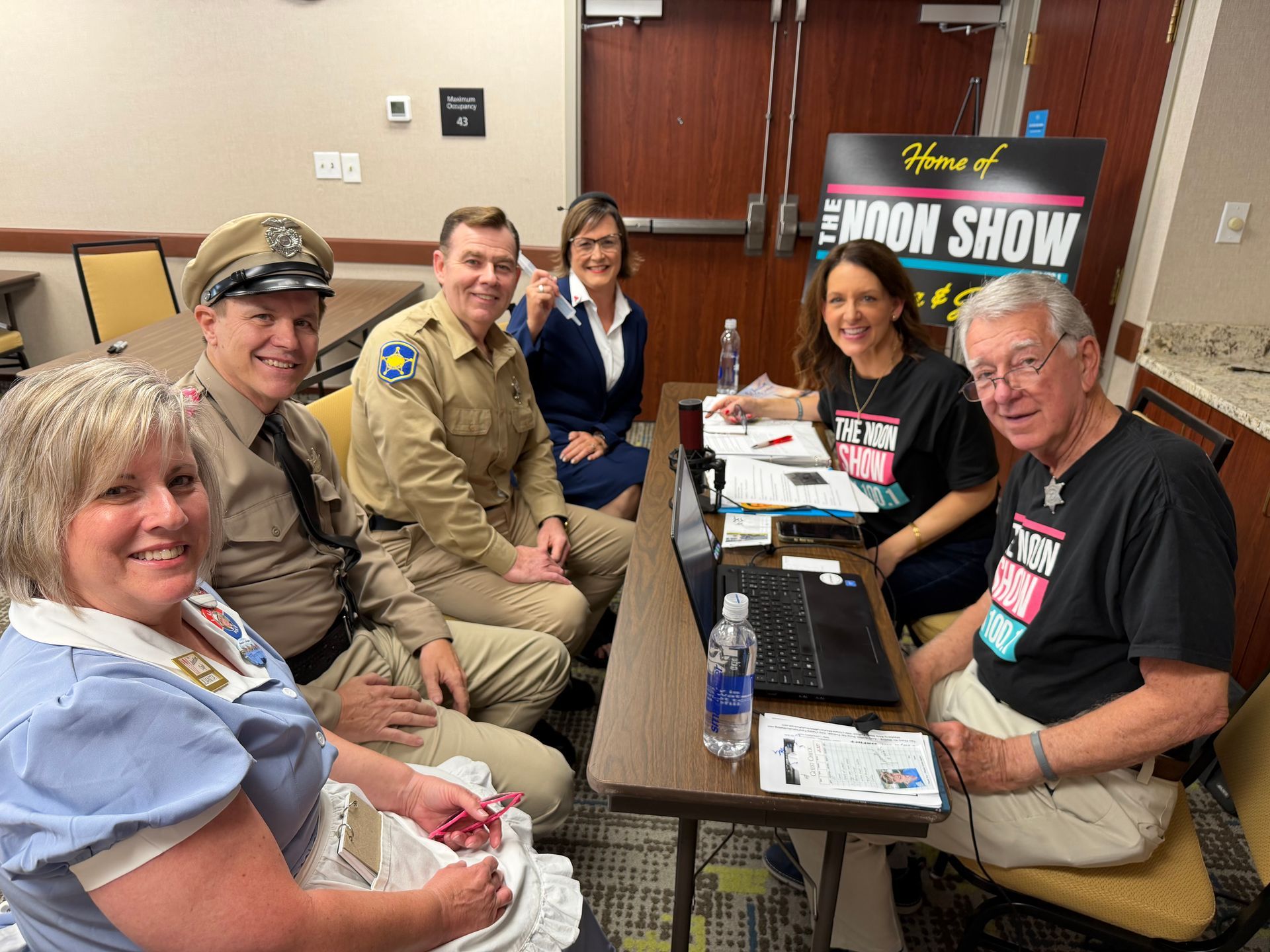 Six people seated at a table in a radio studio, smiling with papers, microphones, and a laptop.