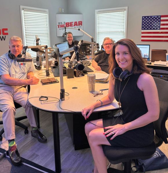 Four people at a radio studio table with microphones, smiling for a podcast recording