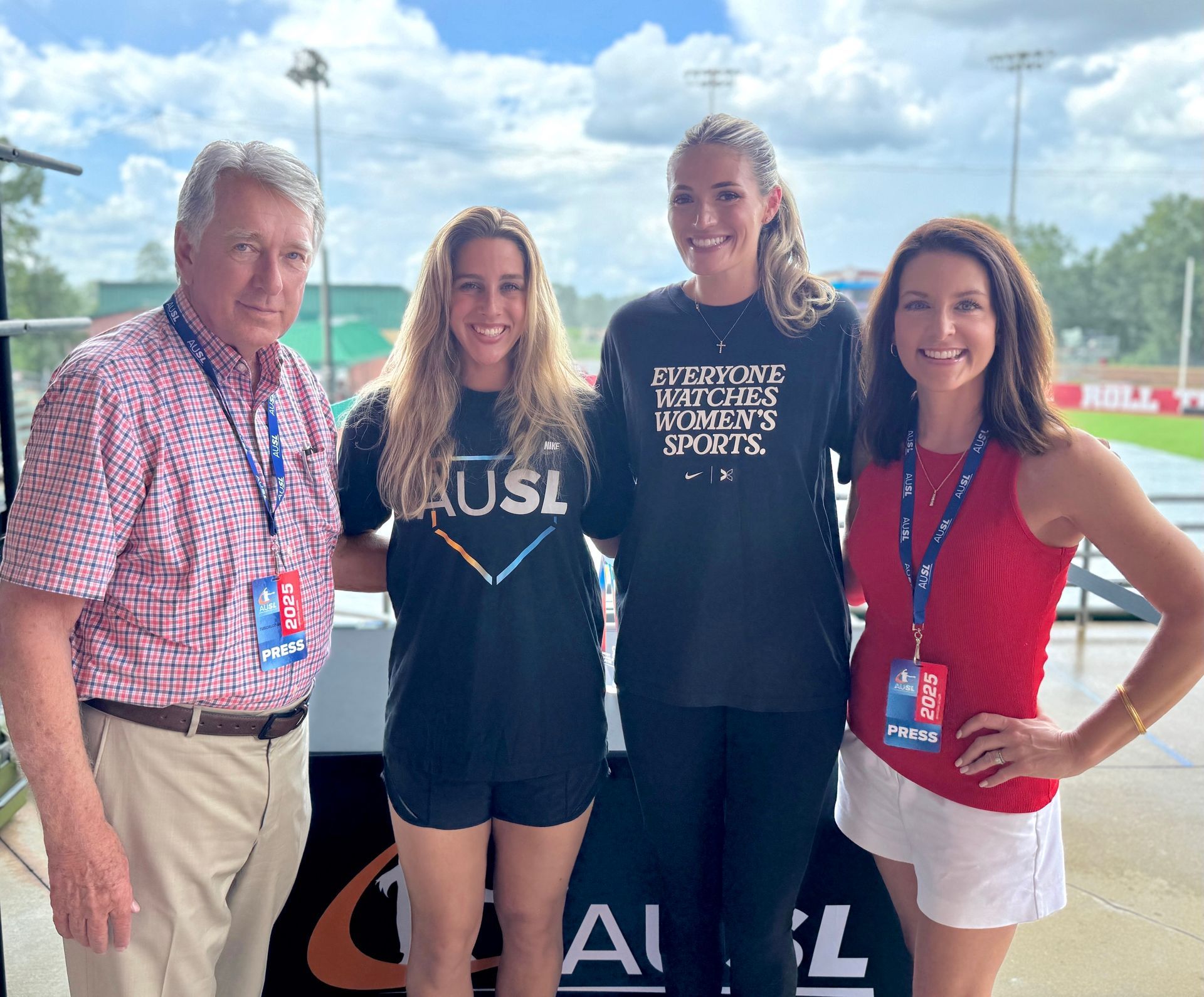Four people posing at a sports venue, with two women in team shirts and a red-shirted woman beside an older man.
