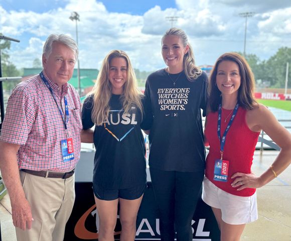 Four people posing at a sports venue, with two women in team shirts and a red-shirted woman beside an older man.