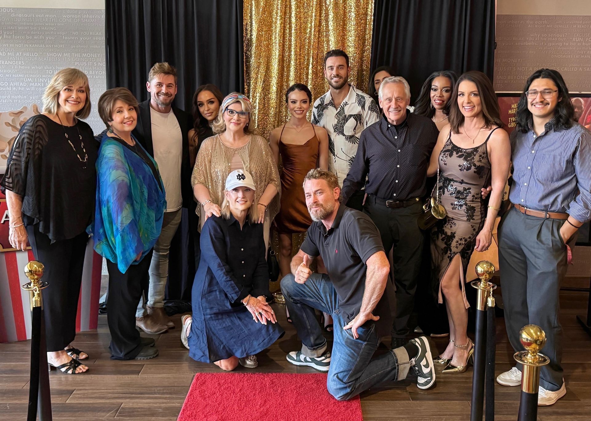 Group posed indoors on a red carpet in front of dark curtains, with two people kneeling in front.