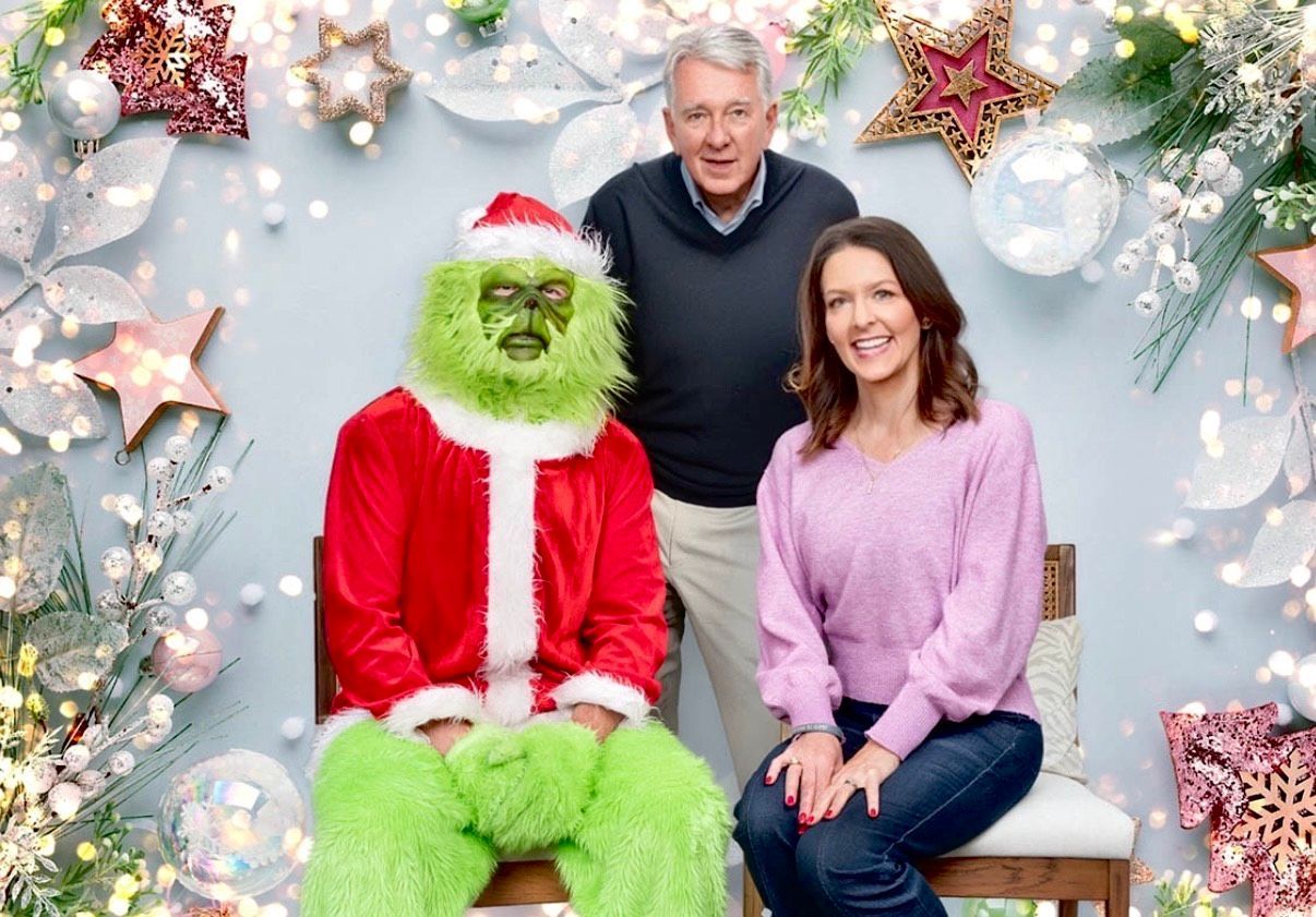 Grinch in Santa suit beside smiling couple in festive holiday backdrop