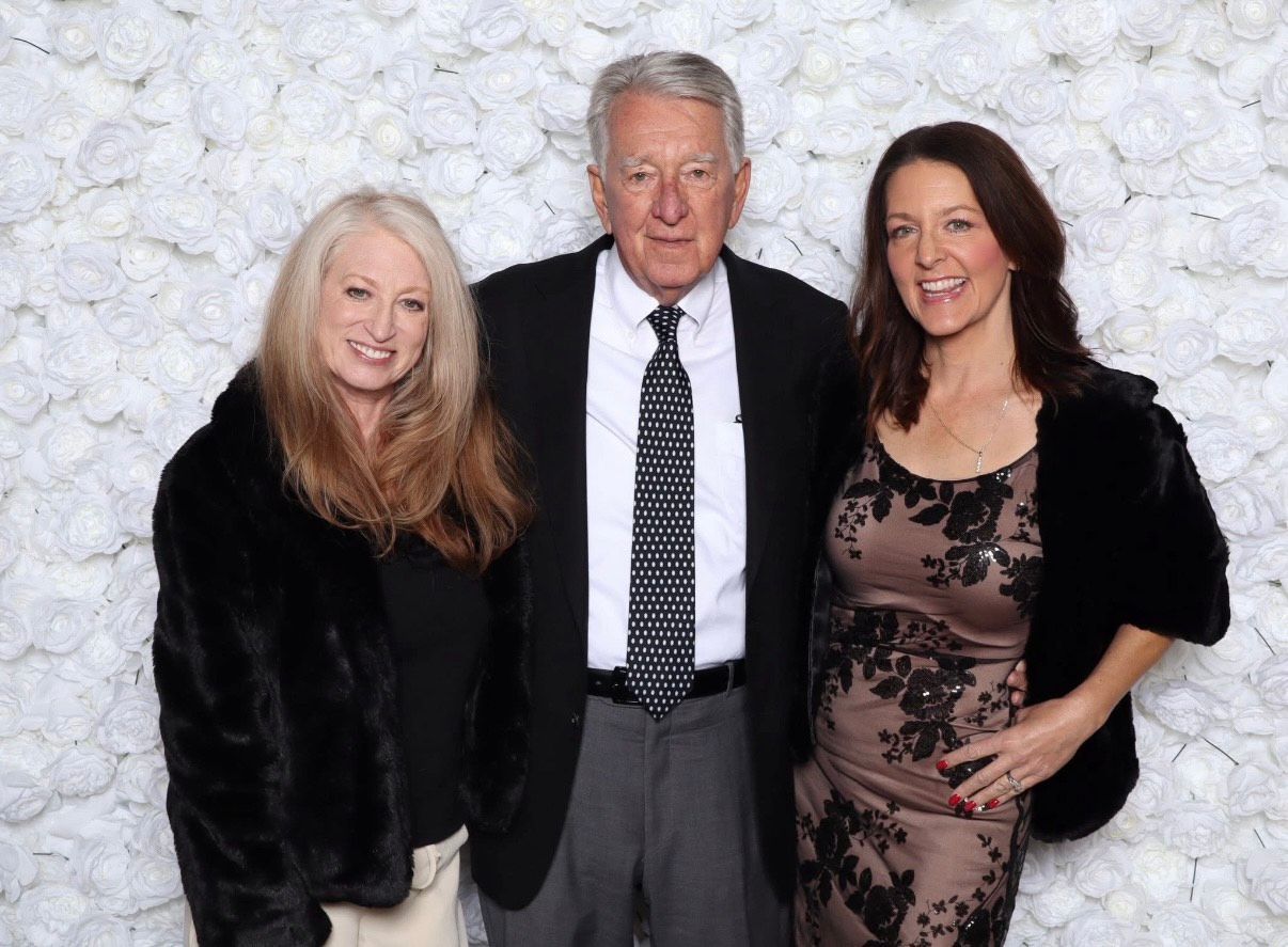 Three people pose together in front of a white stone wall, dressed in formal attire.