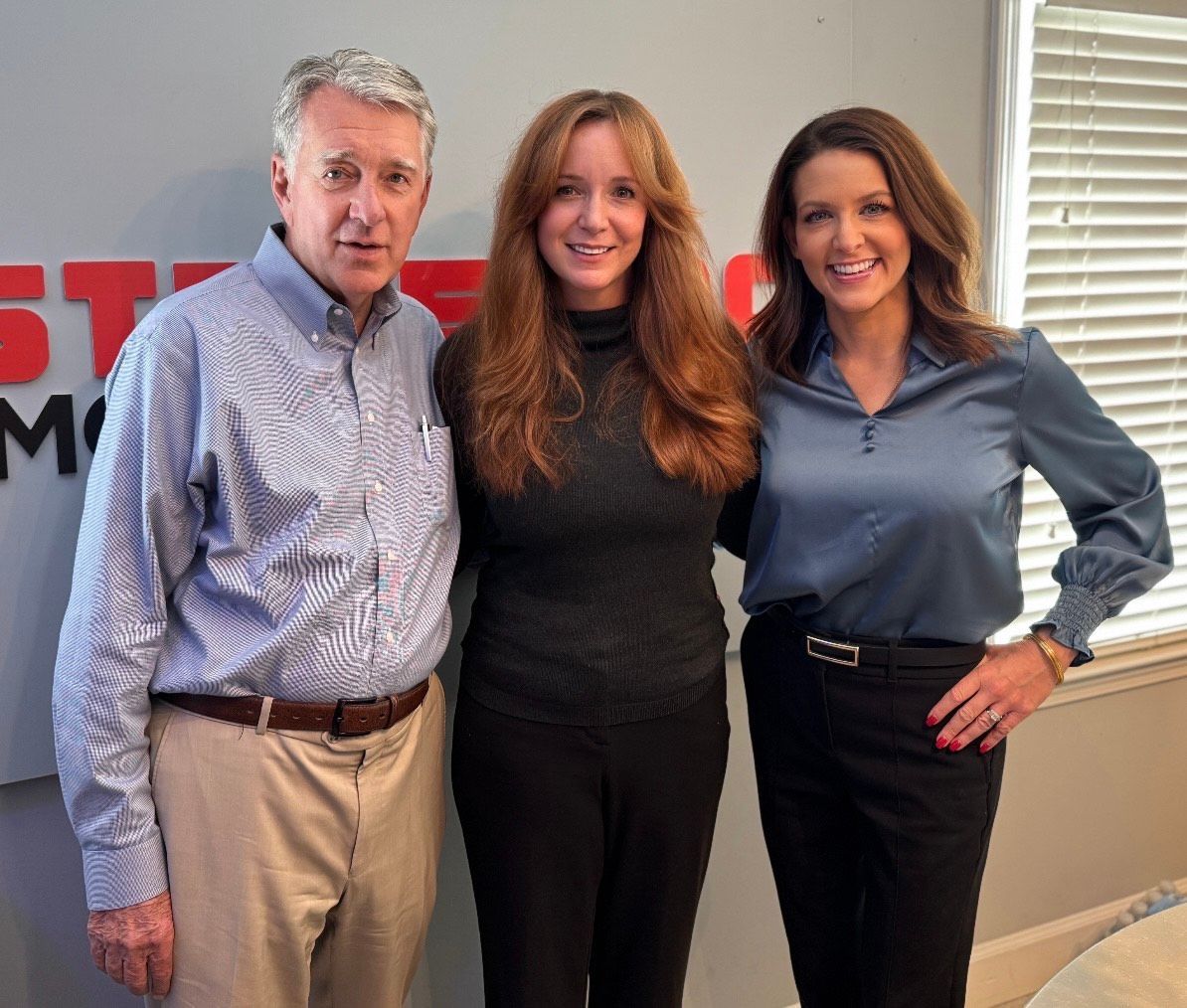 Three people smiling indoors, standing in front of a red and gray office wall logo.