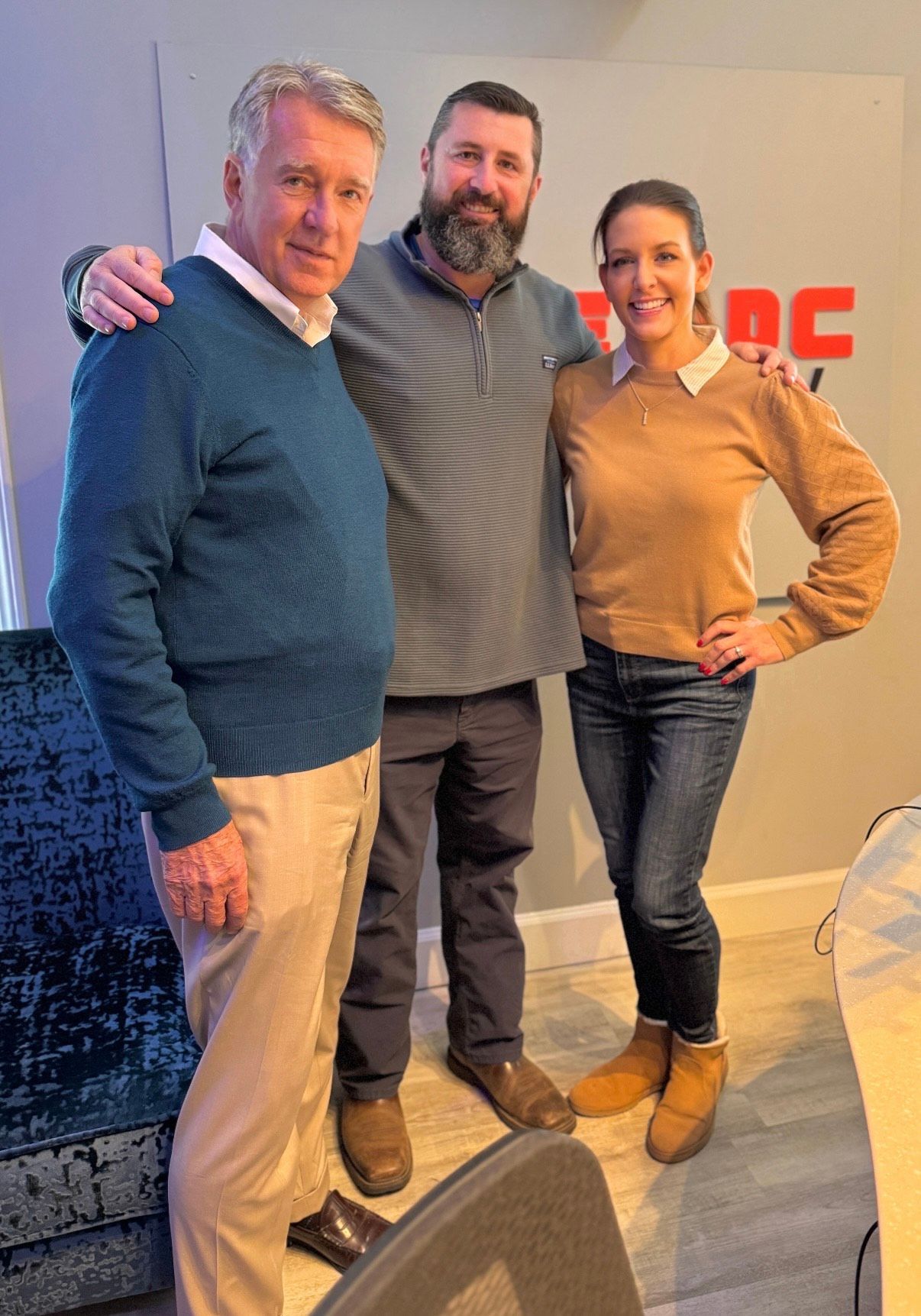 Three adults posing indoors, smiling, in sweaters and jeans against a wall with red lettering.
