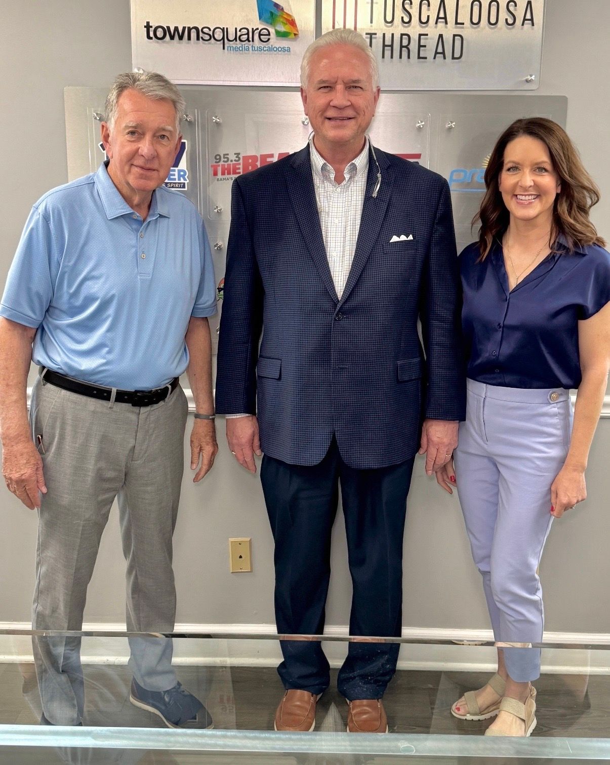 Three people posing indoors before a Tuscaloosa Ahead sign, wearing business casual clothing.
