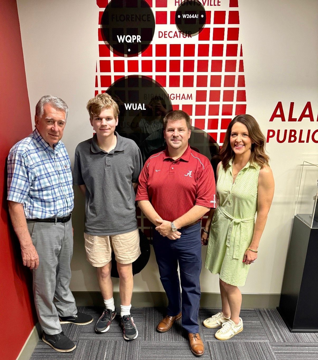 Four people posing in front of a red-and-white wall with public radio station logos.