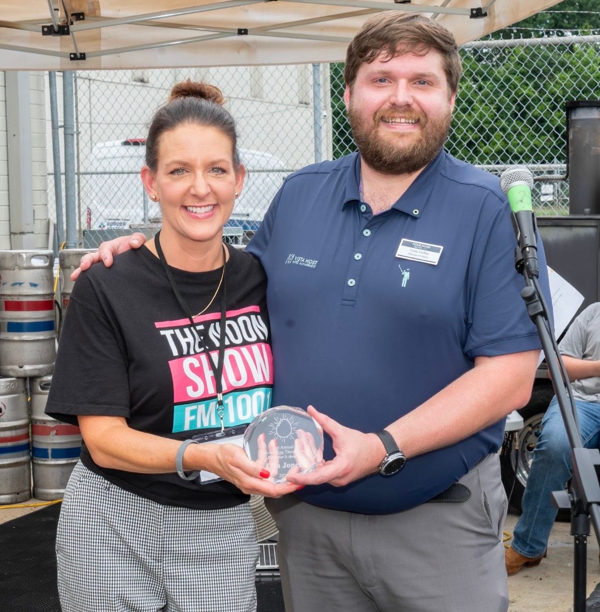 Two people smiling and holding a glass award at an outdoor event booth.