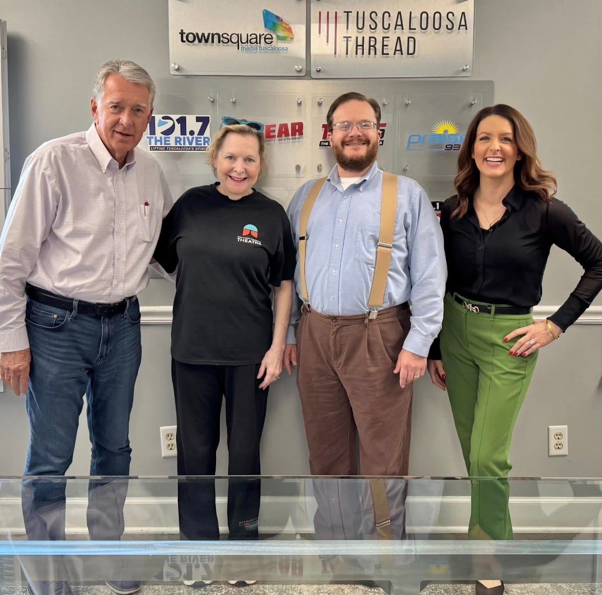Four people standing in a radio studio, smiling in front of station logos.