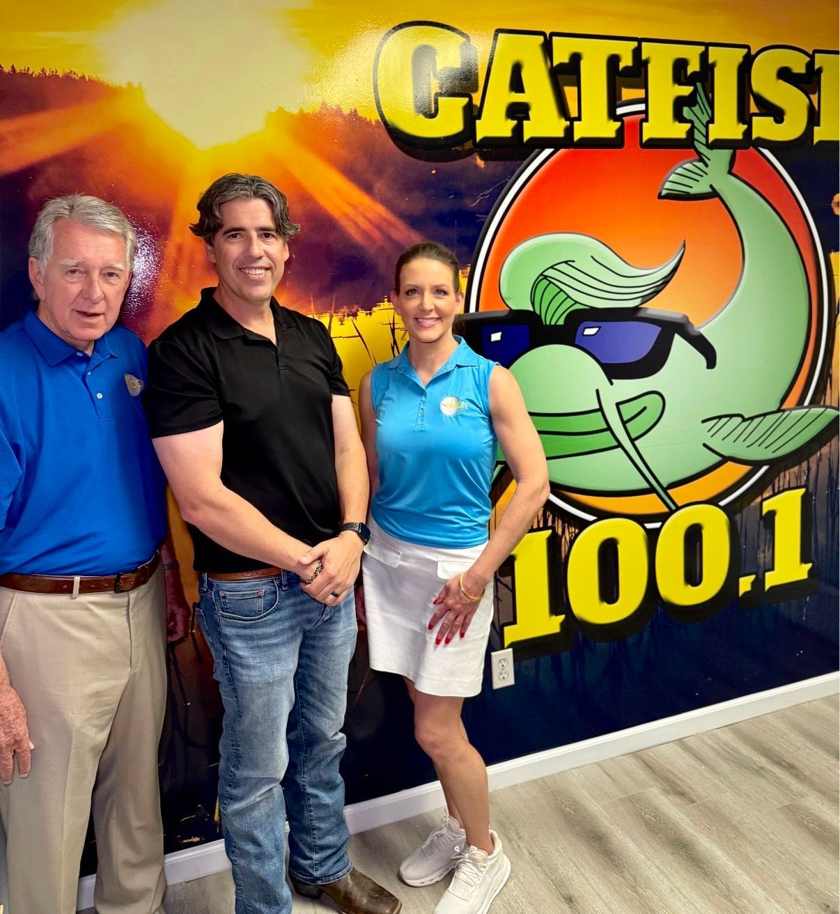 Three people posing in front of a CAT 100.1 radio station logo wall.