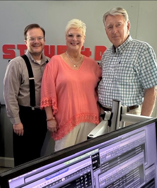 Three people posing in an office studio beside computer monitors and microphones.